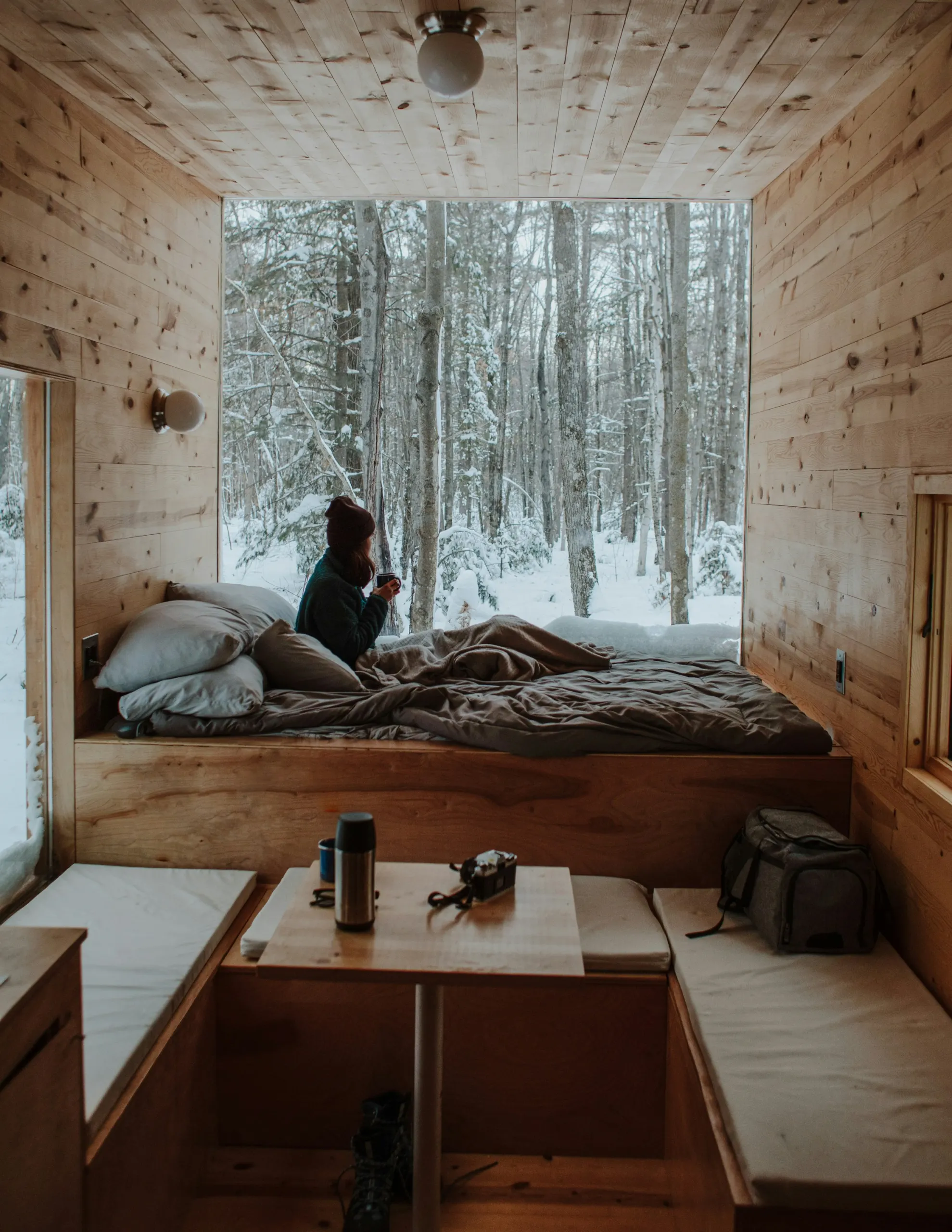 A guest with a hot drink on the bed of a wooden cabin, looking out at a snowy forest