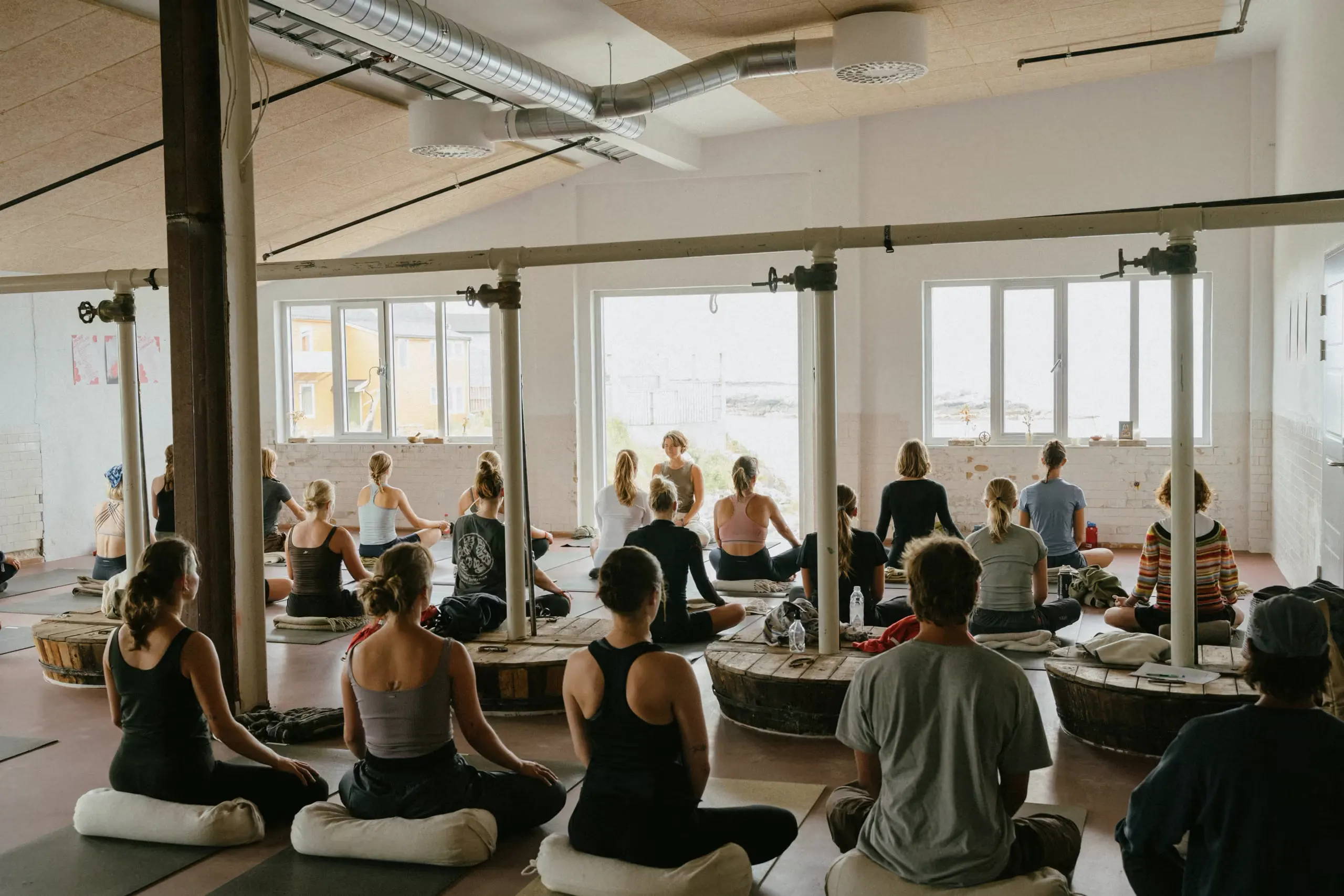A group meditating on cushions in a sunlit hall by the sea